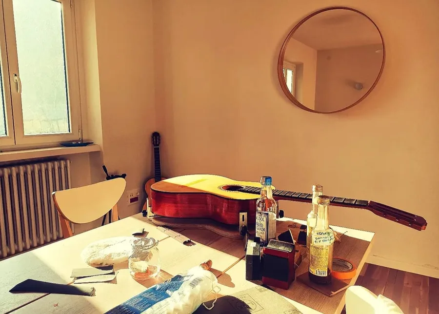 A classical guitar in the white resting on the polishing bench in afternoon light, beside bottles of alcohol and dissolved shellac, a glass jar and cotton pads